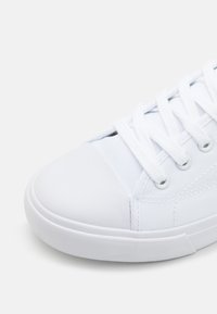 Zapatilla de lona blanca con punta redondeada, protector de goma y parte superior texturizada, con cordones planos y ojales de metal.