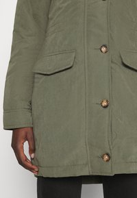 Olivgrön, medellång parka-jacka gjord av texturerat tyg, med två framfickor, knappstängning och synliga sömnadsdetaljer.