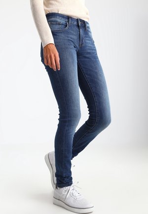Persona che indossa jeans slim fit blu, maglione beige a coste e sneakers bianche, in piedi con un piede leggermente avanti.