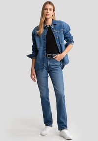 Denim jakna s sprednjim gumbom, dvema žepoma na prsih, oversize kroj. Črna srajca pod njo, v kombinaciji s svetlo modrimi kavbojkami in belimi supergami.