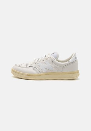 Weißer New Balance Sneaker mit perforierter Spitze, vorderer Schnürung, gepolstertem Kragen und beiger Sohle, nach rechts geneigt auf einfarbigem Hintergrund.