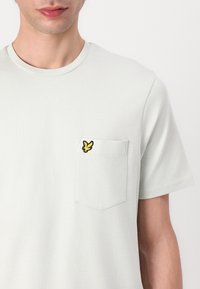 Jasnoszara koszulka z krótkim rękawem, okrągłym dekoltem i kieszonką na lewej piersi, z żółtym haftowanym logo motyla.
