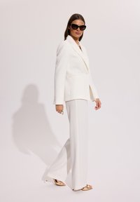 Kobieta w białym, dopasowanym garniturze i złotych szpilkach, nosząca ciemne okulary przeciwsłoneczne, stoi na tle jasnego, jednolitego tła z rozluźnionymi rękami.