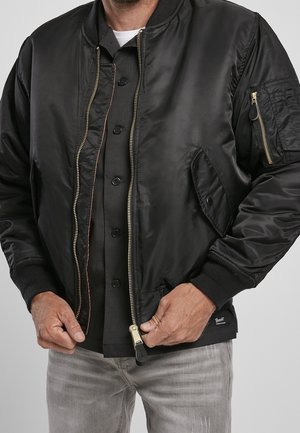 Mężczyzna w czarnej bomberce z mosiężnym zamkiem błyskawicznym, czarnej koszuli na guziki i jasnoszarych dżinsach, ręce regulują zamek kurtki.