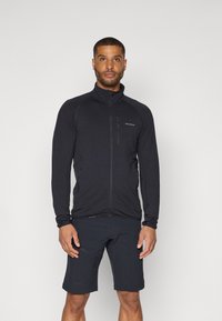 Svart glidelåsjakke laget av teksturert stoff har høy krage, lange ermer og frontlommer med glidelås, sammen med svarte shorts.