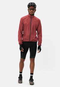 Uomo con una giacca da ciclismo rossa, pantaloni corti imbottiti neri, guanti neri, casco, occhiali da sole e scarpe da ciclismo in piedi su uno sfondo bianco.