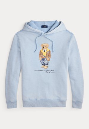 Un hoodie bleu clair avec un ours portant des lunettes, une écharpe jaune, une veste brune, un jean et des baskets, avec le texte "Polo Bear by Ralph Lauren" en dessous.