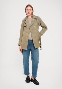 Olijfgroene trenchcoat met een patroon en gouden knopen, gecombineerd met een beige top en lichtblauwe, losse jeans. Zwarte loafers maken de look compleet.