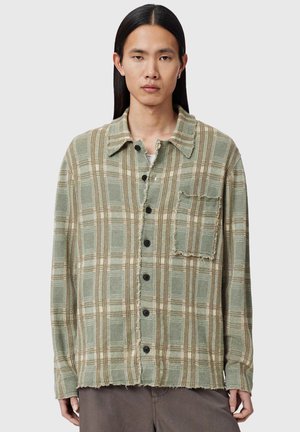 Person iført grøn og beige ternet skjorte med knapper, slidte kanter og lomme med lap, stående foran en ensfarvet lys baggrund.