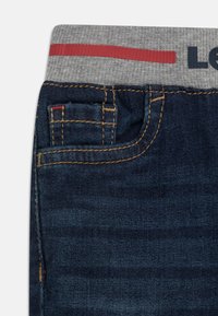 Jean en denim avec une taille grise présentant une bande rouge. Poche avant courte, couture contrastante et une texture légèrement délavée.