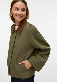 Olivno zelena jakna s rjavim ovratnikom, zadrgo, stranskimi žepi in gumbki na ramilih. Ima sproščen kroj in gladko teksturo.