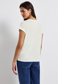 Short-sleeve, cream-colored top with soft texture, rounded neckline, and rolled cuffs, paired with dark blue jeans.
