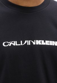 Nærbillede af sort Calvin Klein t-shirt med hvidt logotekst på tværs af brystet, der viser halsudskæring og den øverste del af bæreren.
