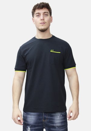 Giovane uomo che indossa una maglietta nera con rifiniture giallo neon sulle maniche e sulla tasca, abbinata a jeans blu strappati, in piedi in posizione neutra.