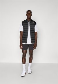 Gilet matelassé noir sur un t-shirt blanc, assorti à un short gris. Le mannequin porte des baskets blanches avec des accents noirs et des chaussettes mi-mollet.