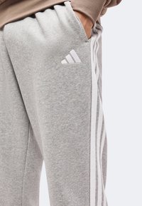 Άτομο που φοράει γκρι φόρμα Adidas με λευκές ρίγες στο πλάι και κεντημένο λογότυπο Adidas κοντά στην τσέπη με το χέρι μέσα.