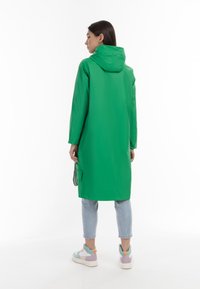 Cappotto impermeabile verde con cappuccio, maniche lunghe e vestibilità rilassata. Presenta una tasca sul retro ed è abbinato a jeans corti blu chiaro e sneakers colorate.