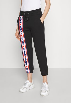 Osoba ubrana w czarne joggery z białym, czerwonym i niebieskim paskiem bocznym z napisem „GCDS WEAR” oraz białe trampki, stojąca na jednolitym tle.