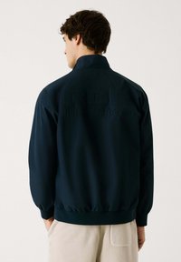 Veste bomber bleu marine avec col haut, poignets élastiques et ourlet. Présente le logo en relief "RBR RED BULL RACING" au dos. Texture lisse.