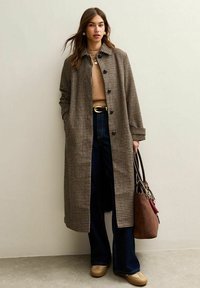 Abrigo de pata de gallo en marrón y crema, corte ajustado, mangas largas, combinado con un suéter beige, jeans oscuros de pierna ancha y un bolso marrón.
