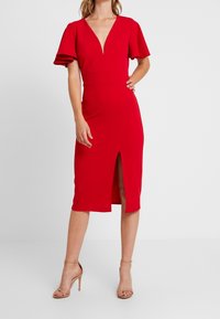 Czerwona sukienka z głębokim dekoltem w literę V, krótkimi, falbankowymi rękawami oraz rozcięciem z przodu. Stylizacja jest dopasowana do sylwetki i uszyta z gładkiego, elastycznego materiału.