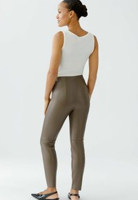 Femme avec les cheveux en chignon portant un haut blanc sans manches, un pantalon en cuir taupe taille haute et des chaussures à talon noir, se tenant de profil sur fond blanc.