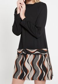 Svart långärmad topp som är kombinerad med ett mönstrat kjol med vågiga ränder i beige, svart och rostfärg, accentuerad med en svart bältesspänne.