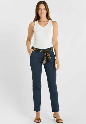 Vrouw draagt een witte mouwloze top, marineblauwe broek met een luipaardprintriem en bruine open-toe hakken, staand tegen een effen lichte achtergrond.