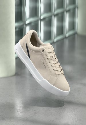 Beiger Sneaker mit niedrigem Schaft, weißer Sohle, Schnürsenkeln und dezenter Markenkennzeichnung, schwebend über einem polierten Betonboden nahe einer Glasbausteinwand präsentiert.