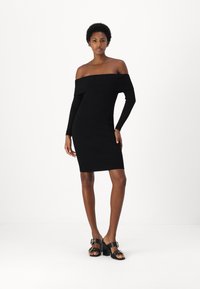 Abito nero a coste off-shoulder con maniche lunghe, forma aderente, lunghezza al ginocchio. Abbinato a sandali neri con tacco che presentano dettagli dorati.