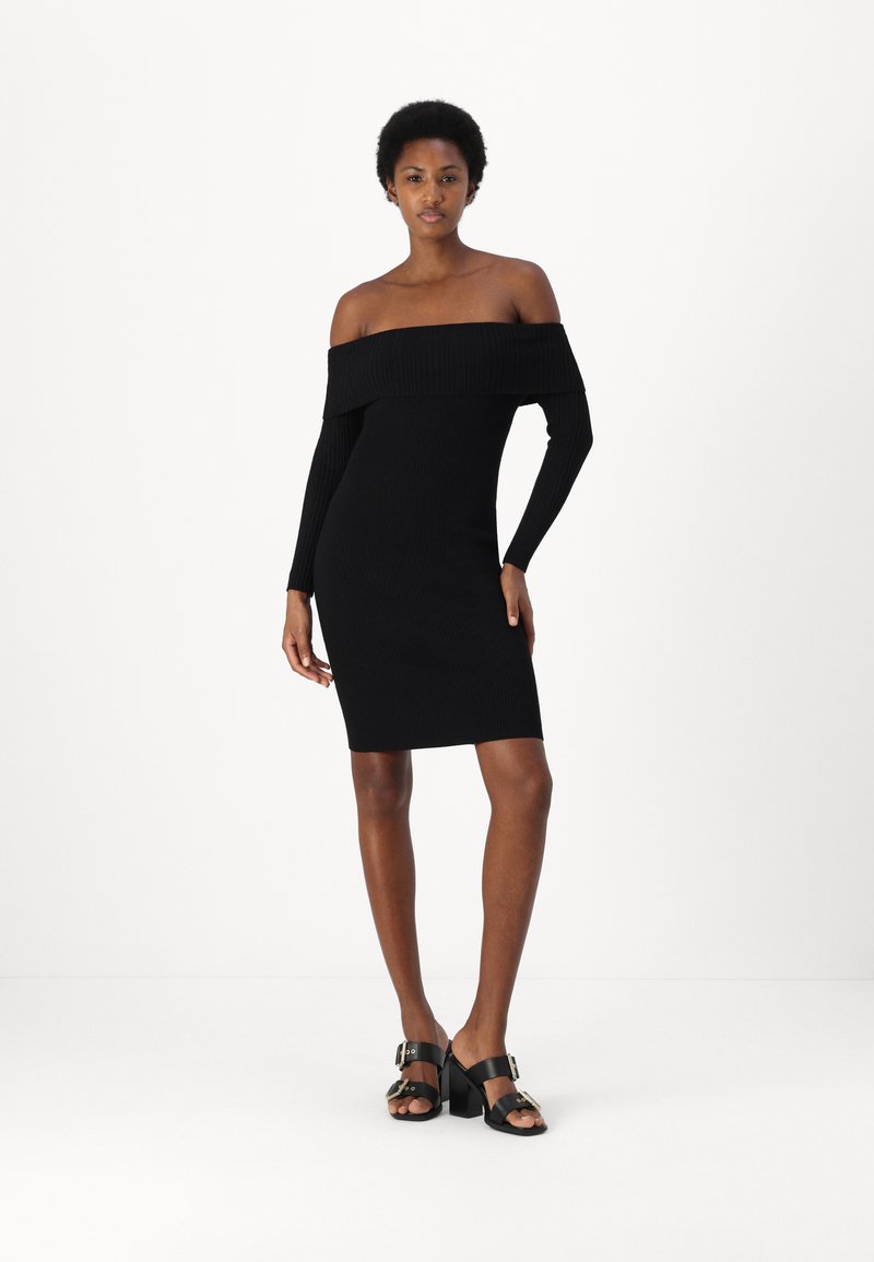 Abito nero a coste off-shoulder con maniche lunghe, forma aderente, lunghezza al ginocchio. Abbinato a sandali neri con tacco che presentano dettagli dorati.