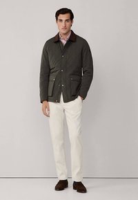 Giacca trapuntata verde scuro con colletto marrone, con due tasche frontali, indossata sopra una camicia a righe e pantaloni bianchi.