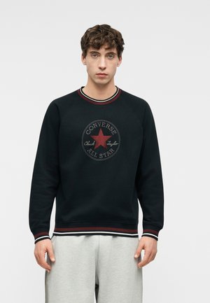 Mladý muž oblečený v čiernej mikine Converse All Star s červeným hviezdičkovým logom a svetlosivých teplákoch, stojaci s neutrálnym výrazom.