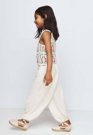 Jeune fille en haut sans manches à motifs, pantalon blanc ample et espadrilles beige marchant de côté sur un fond uni.