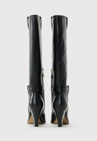 Pair of knee-high black leather heeled boots with rear zipper and strap detail against white background.