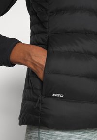 Svart isolerad jacka med quiltat mönster, som har en sidoficka och en detalj med "550"-logotyp. Materialet verkar slätt och lättviktigt.