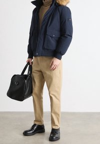 Marineblauwe pufferjack met een met bont gevoerde capuchon, voorzien van twee voorzakken. Lichtbruine broek en zwarte schoenen, met een zwarte sporttas.