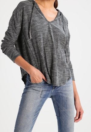 Kvinde iført en løs, struktureret mørkegrå langærmet top med kvastdetaljer, kombineret med lyseblå denimjeans med synlige lapper.