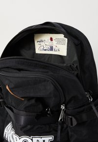 Sac à dos noir avec une surface texturée, doté de multiples zips, d'une étiquette intérieure avec des illustrations et de bretelles ajustables.