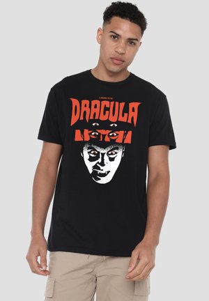 Young man wearing a black T-shirt with red text "Dracula" and a white graphic of a vampire face with multiple eyes on a plain light background.