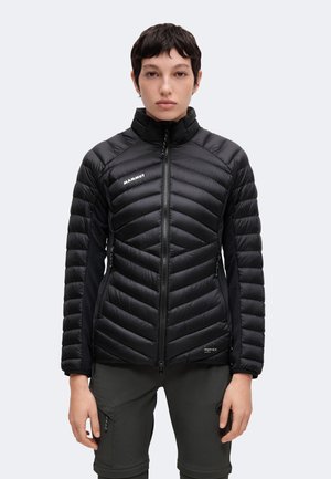 Persona que lleva una chaqueta acolchada negra de Mammut con cremallera frontal y pantalones oscuros, de pie frente a un fondo liso claro.