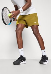 Nike τένις σόρτς σε ελαιόμαυρο, με λευκό λογότυπο. Μια λευκή μπλούζα, περιδέρο χεριού και μαύρα αθλητικά παπούτσια με λευκές λεπτομέρειες. Κρατώντας μια μπάλα τένις και ρακέτα.