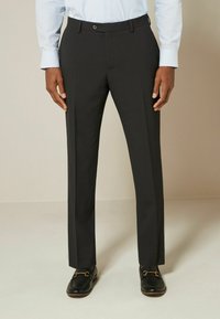 Pantalons tailleur gris foncé avec une coupe plate, ajustée, et des détails de pli, associés à des mocassins noirs avec des accessoires décoratifs.