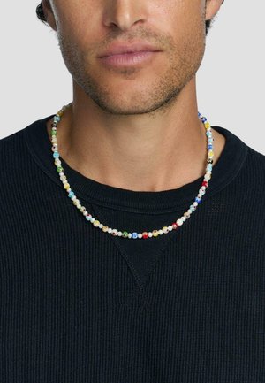 Mand iført en flerfarvet perlekæde og en sort ribbet crew neck sweater med lidt ansigtshår synligt.