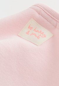 Tissu rose clair avec une étiquette rectangulaire blanche cousue portant la mention « be happy & smile » en texte cursif rose près d'un bord côtelé.