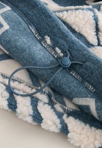 Sininen fleecytyyny, jossa on geometrinen kuvio kermavärisenä, narukiristys ja tekstuuriset reunat lisäyksinä.