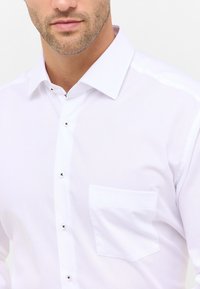 Camicia bianca con colletto a punta, maniche lunghe e una tasca piatta. Tessuto liscio con una leggera lucentezza e bottoni argentati.