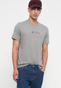 Γκρι βαμβακερό μπλουζάκι με κοντά μανίκια και στρογγυλή λαιμόκοψη, που διαθέτει λογότυπο σε μπλε, κόκκινο και μαύρο χρώμα. Συνδυάζεται με μπλε τζιν και μπορντό καπέλο.