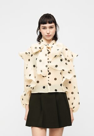 Młoda kobieta w przezroczystej kremowej bluzce w czarne groszki, z dużą kokardą, falbankowymi rękawami i czarnej plisowanej spódnicy.