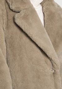 Nærbillede af beige imiteret pelskåpekrave over hvid ribstrikket rullekrave, der viser tekstur og knaphuldsdetalje.